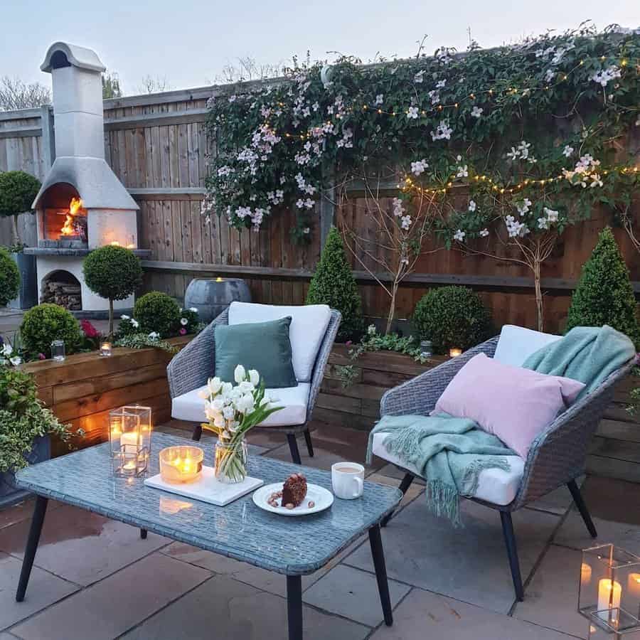 Cozy outdoor patio with two cushioned chairs, a table with candles and cake, a lit fireplace, and plants with string lights on a fence