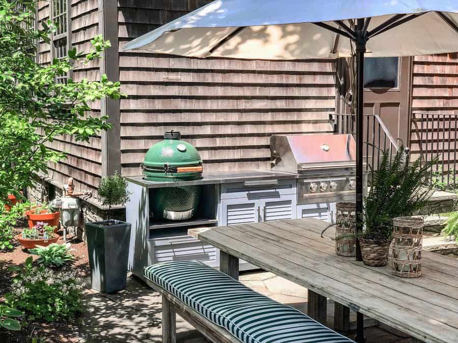 Patio with a wooden table, striped bench, grill, and green ceramic cooker under a large umbrella, surrounded by greenery