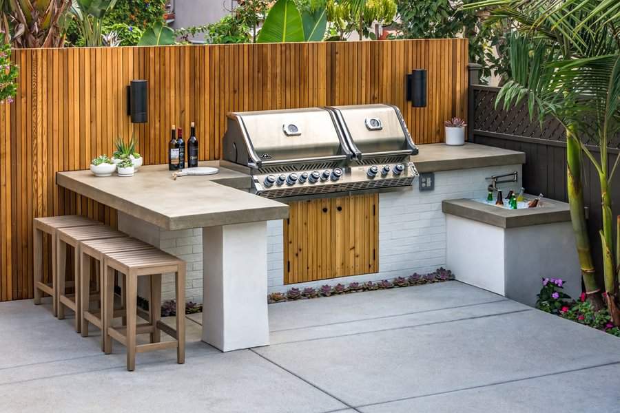 Outdoor kitchen with a stainless steel grill, concrete countertop, wooden stools, and potted plants, set against a wooden paneled wall