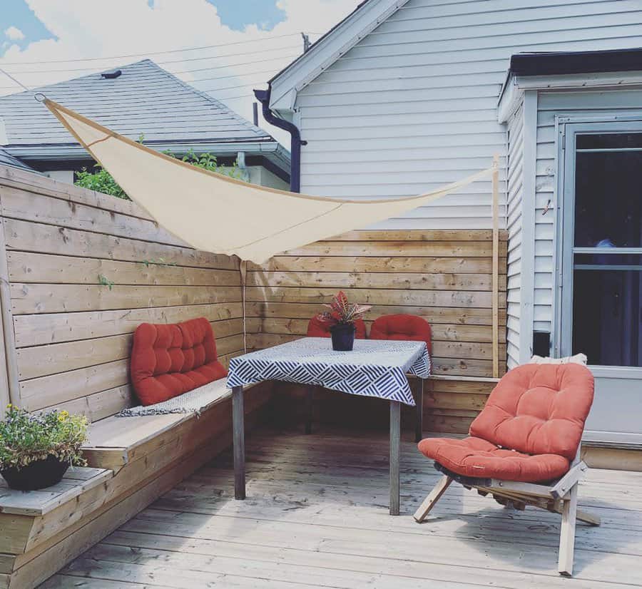 Small patio with wooden benches, red cushions, a table with a patterned cloth, potted plant, and a sunshade canopy