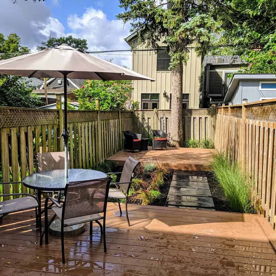 Wooden deck with a round table and umbrella; pathway leads to a seating area with chairs; tall fence and tree in the background