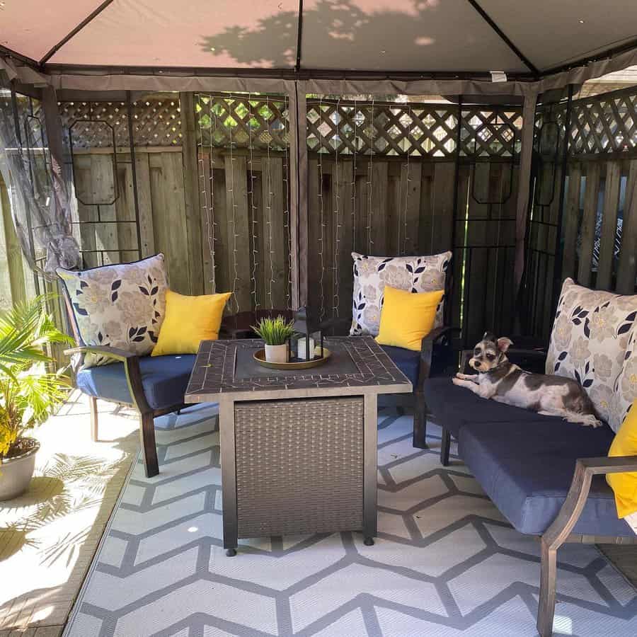 Cozy gazebo-covered patio with cushioned seating, yellow accent pillows, a fire pit table, string lights, and a relaxed dog on the bench