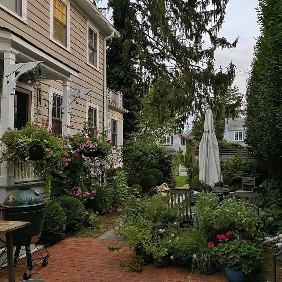 Cozy garden patio with a brick floor, surrounded by lush plants and flowers, next to a charming two-story house, umbrella and chairs visible