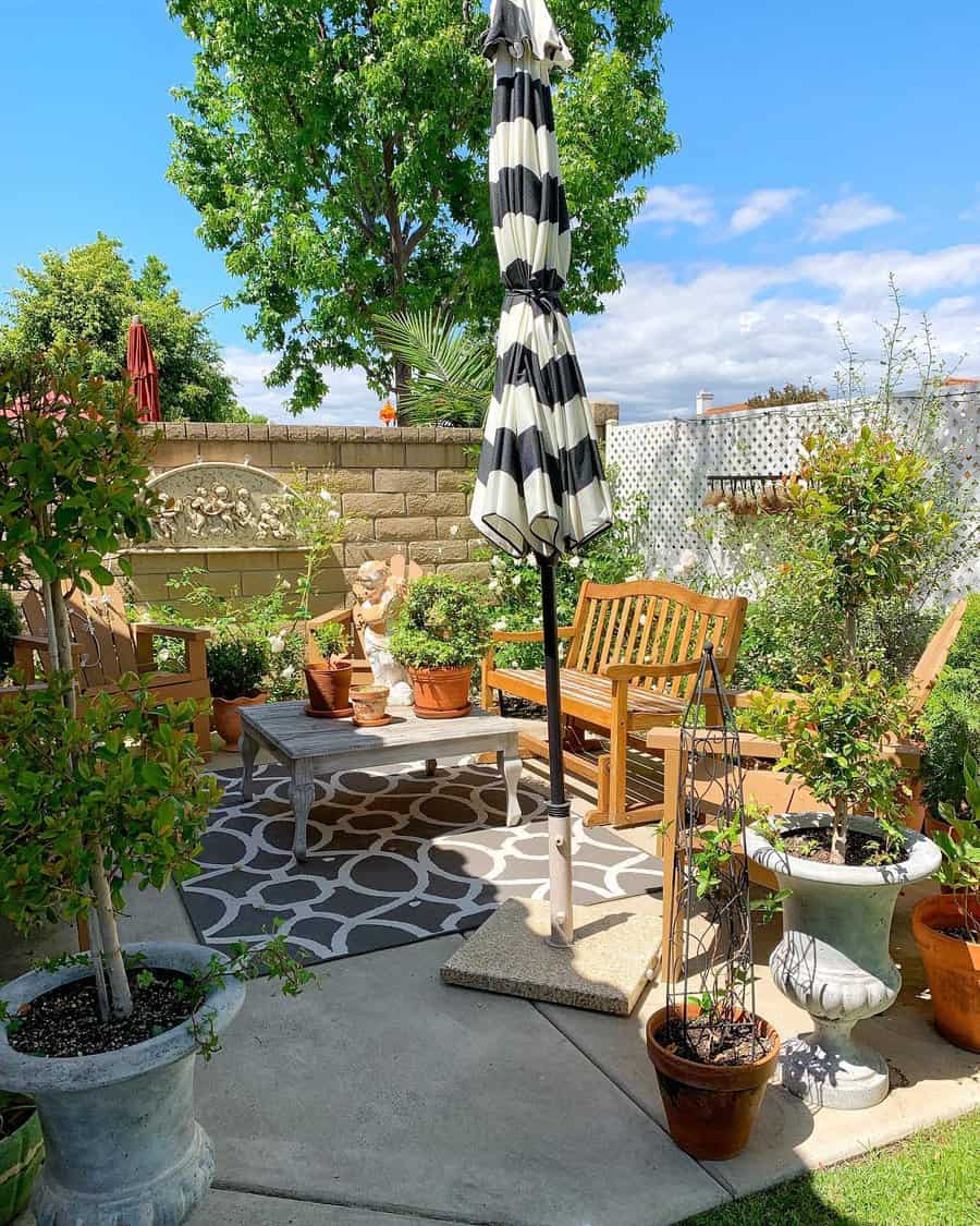 Sunny garden patio with a wooden bench, striped umbrella, potted plants, and a small fountain; trees and a lattice fence surround the area