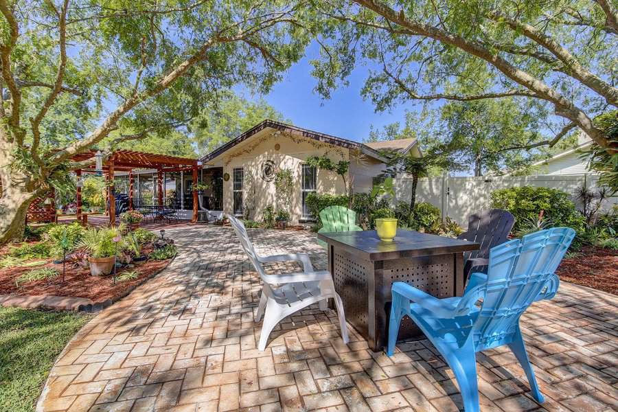 Backyard with brick patio, tree shade, seating area with colorful adirondack chairs, and a small table, bordered by plants and a wooden pergola