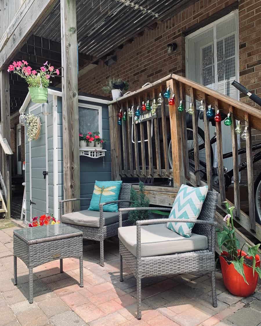 Cozy outdoor patio with wicker chairs, a small table, and potted plants; bright string lights and decor adorn the wooden railing