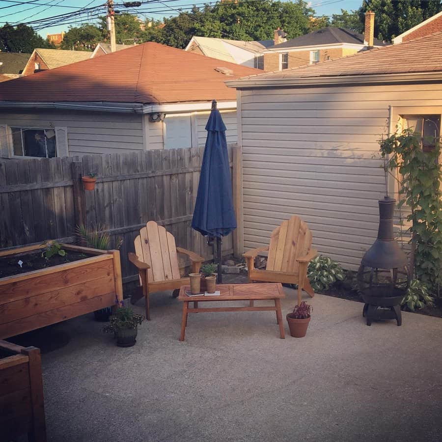 Cozy backyard patio with wooden adirondack chairs, a bench, and a table with a blue umbrella, complemented by plants and a chiminea decorating the space