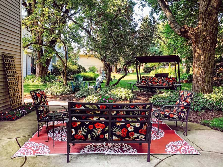 Backyard patio with floral cushioned furniture on a red-patterned rug, surrounded by lush trees and greenery