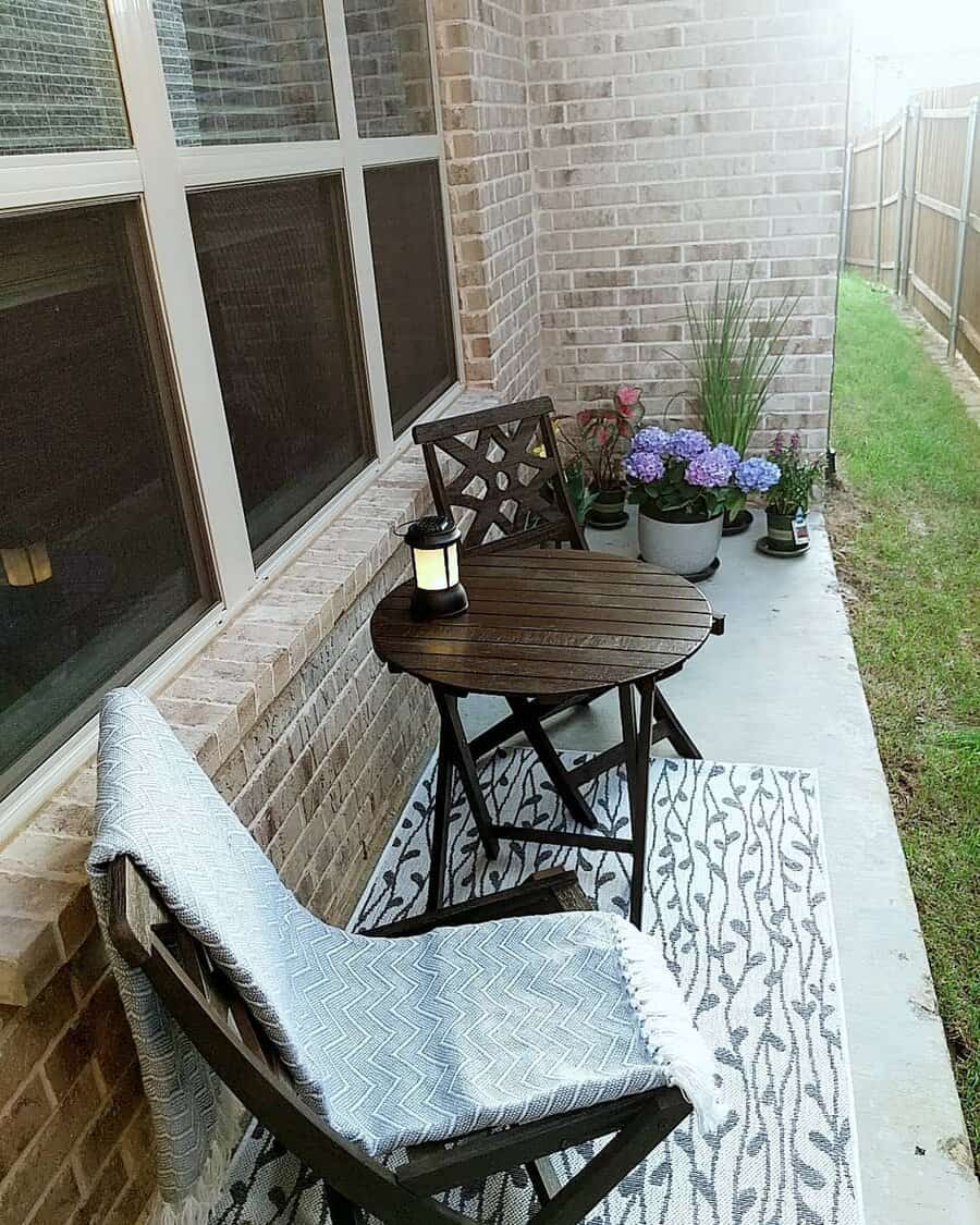 Cozy patio with a wooden table and chair, a blanket, a candle lantern, potted plants, and a brick wall backdrop