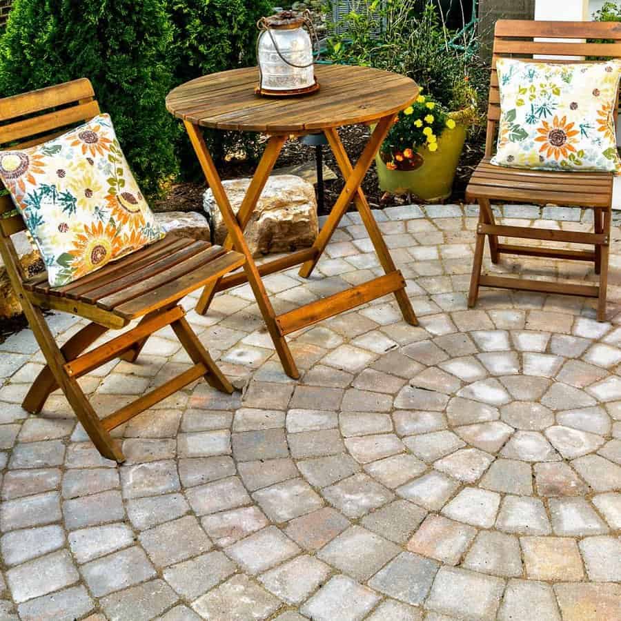 Wooden patio set with floral cushions on a circular stone pattern, featuring a lantern centerpiece, with green shrubs and yellow flowers in the background