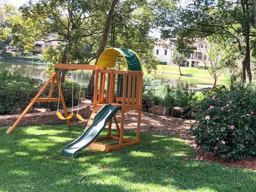 Playground set with a slide, swing, and climbing area on lush grass near a serene lake, surrounded by trees and shrubs