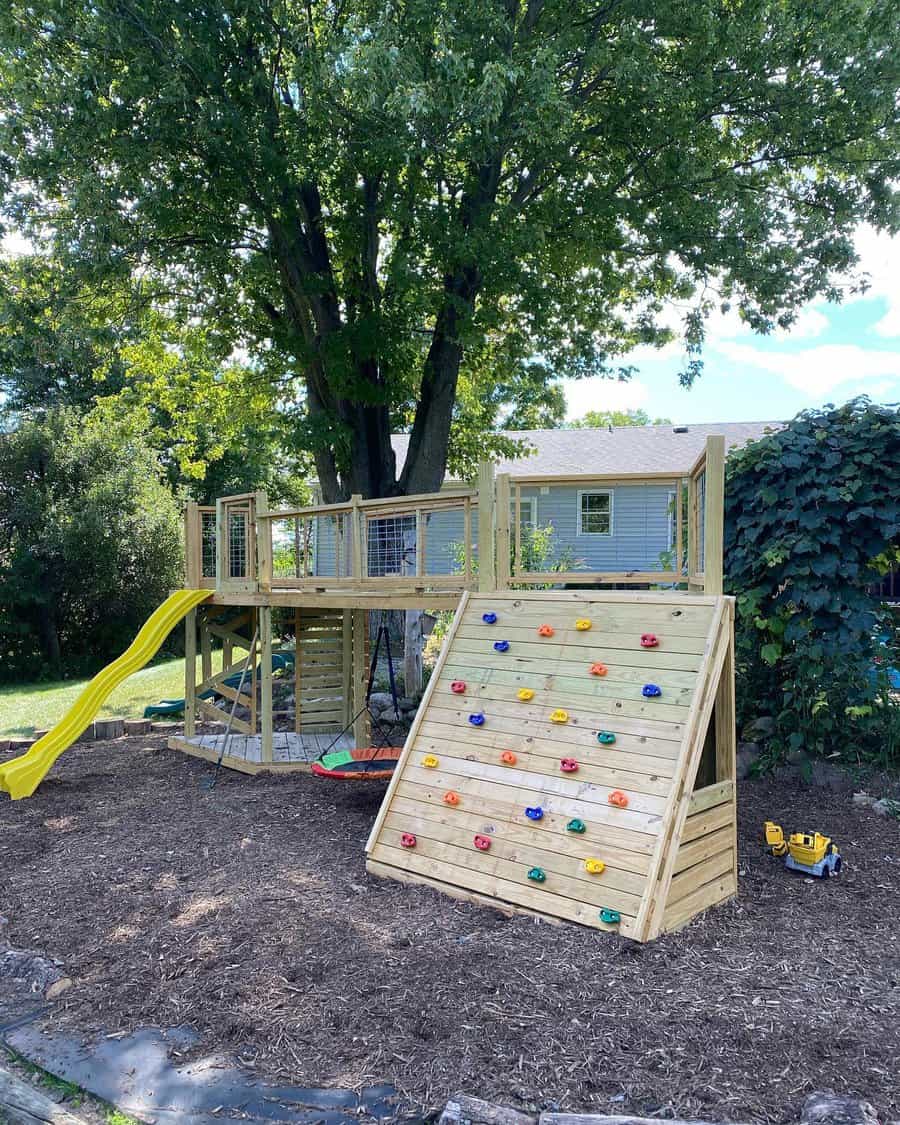 Wooden backyard playground with a yellow slide, climbing wall, and elevated play deck under a large tree, creating a fun outdoor space