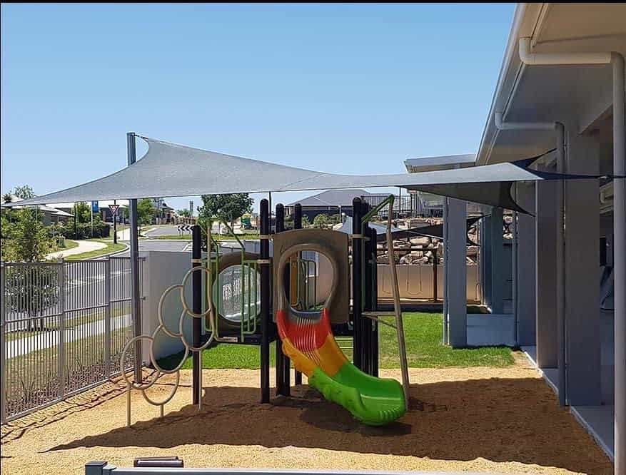 Playground with a modern shade sail providing sun protection, featuring a colorful slide and climbing elements on a soft surface