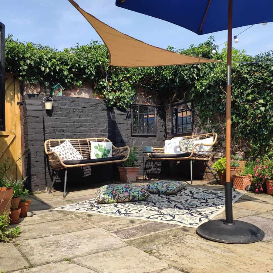 Cozy backyard patio with a shade sail and umbrella, featuring rattan benches, potted plants, and a patterned outdoor rug