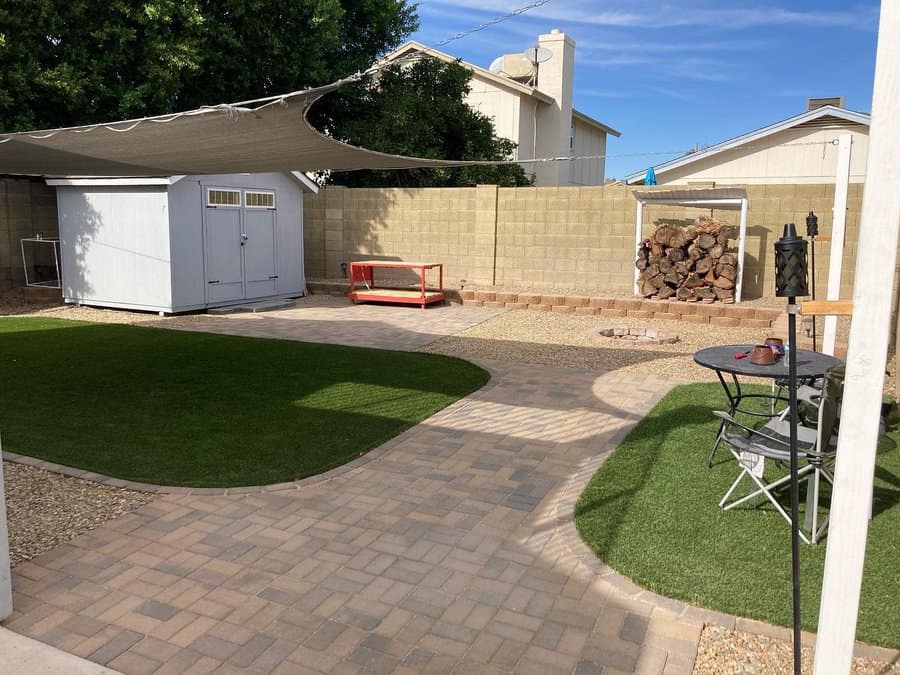 Backyard with a large shade sail, artificial grass, a firewood stack, a fire pit, and a cozy seating area on a paved patio