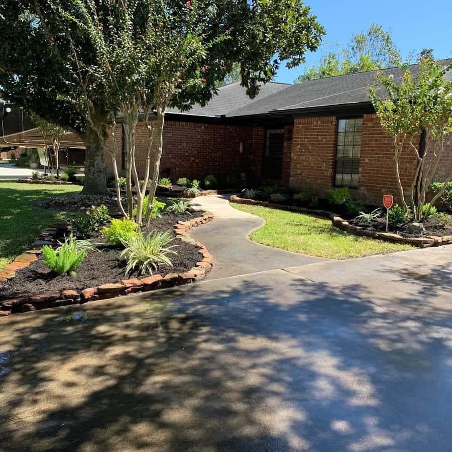 Front yard landscaping with a curved walkway, brick edging, fresh mulch, vibrant plants, and shady trees leading to a brick home