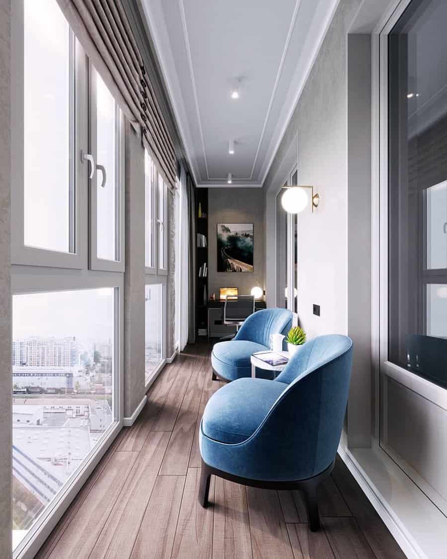 Elegant enclosed balcony with floor-to-ceiling windows, plush blue chairs, and a cozy reading nook, offering a relaxing city view