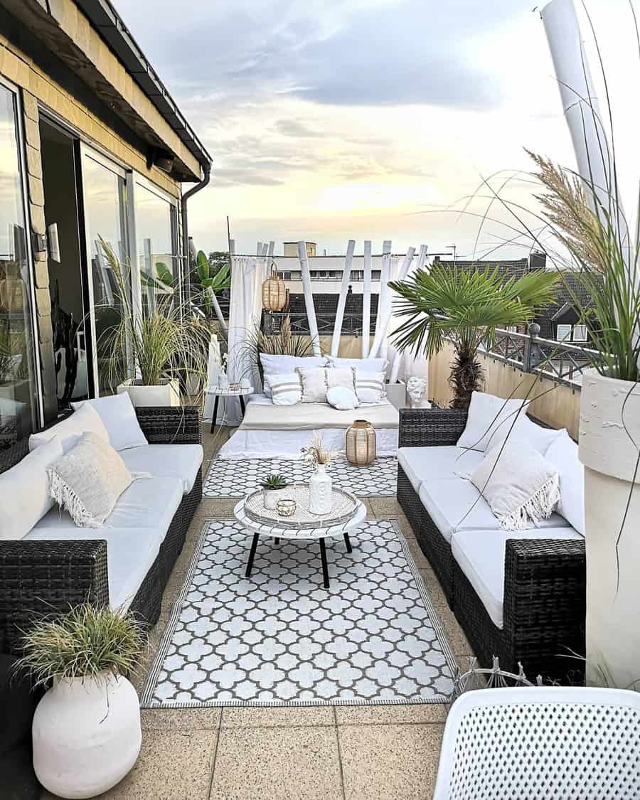 Rooftop terrace with wicker sofas, white cushions, patterned rug, coffee table, and potted plants under a cloudy sky