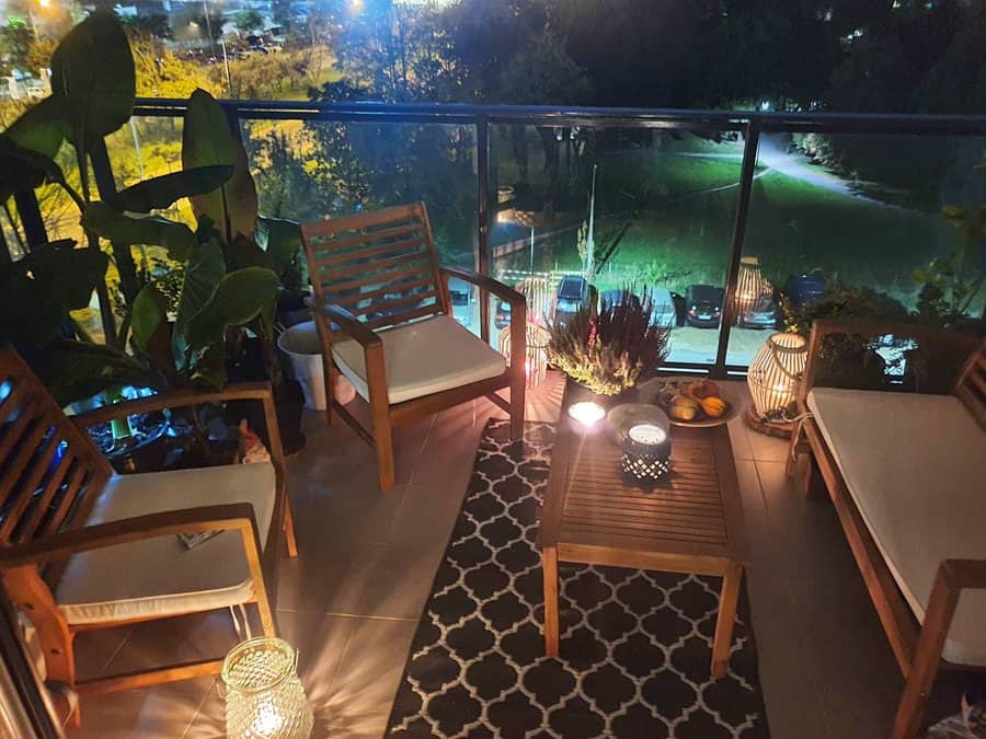 Cozy balcony with wooden furniture, cushions, lanterns, and plants; night view of trees and city lights in the background