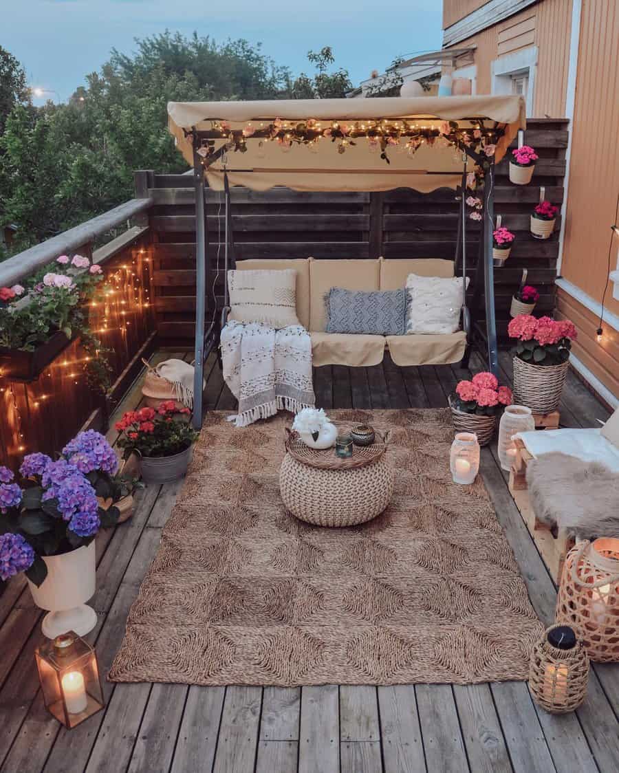 Cozy balcony with a swing sofa, cushions, fairy lights, flowers, candles, and a woven rug creating a warm and inviting ambiance