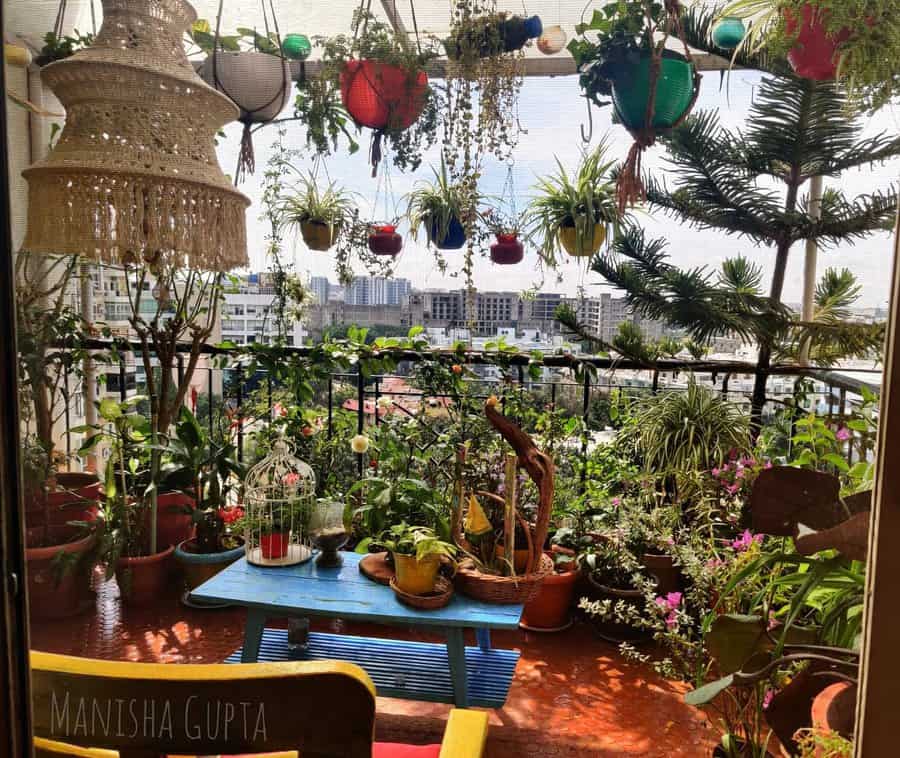 Lush balcony garden with colorful hanging planters, potted greenery, a blue wooden table, and a macramé lamp, overlooking the city