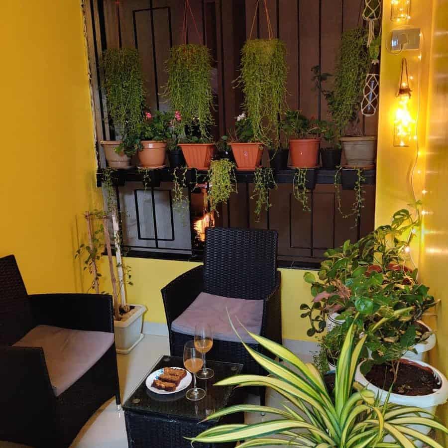 Cozy balcony with wicker chairs, table with snacks and drinks, potted plants on railing, and warm string lights on yellow wall