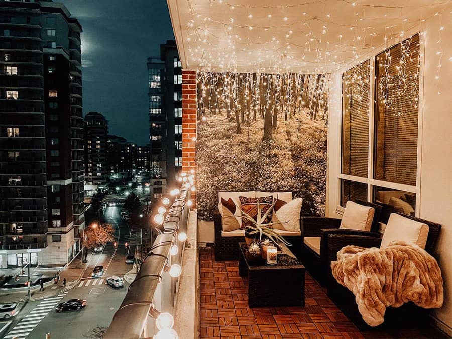 Cozy balcony with string lights, wicker chairs, and a throw blanket; tapestry with forest scene on wall; city view at night