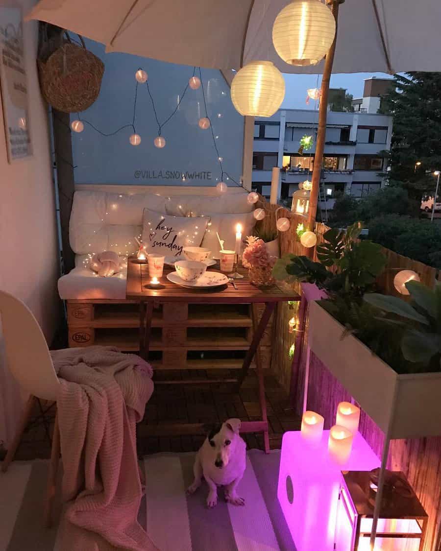 Cozy small balcony at night with string lights, candles, a pallet sofa, a wooden dining table, lanterns, and a small dog