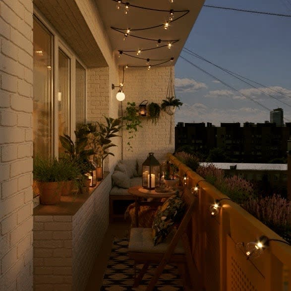 Cozy balcony at night with warm string lights, potted plants, a wooden table with lanterns, and a cushioned seating nook