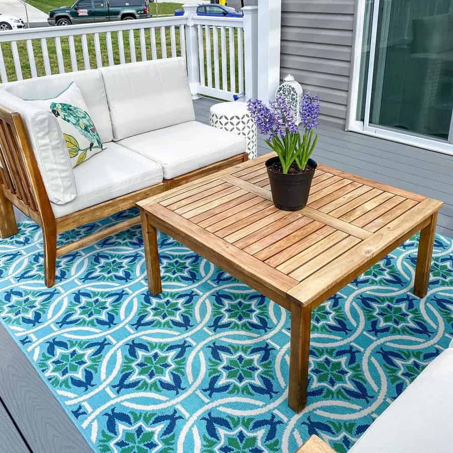 Outdoor patio with wooden furniture, white cushions, and a floral pot on a wooden table, blue patterned rug on the floor