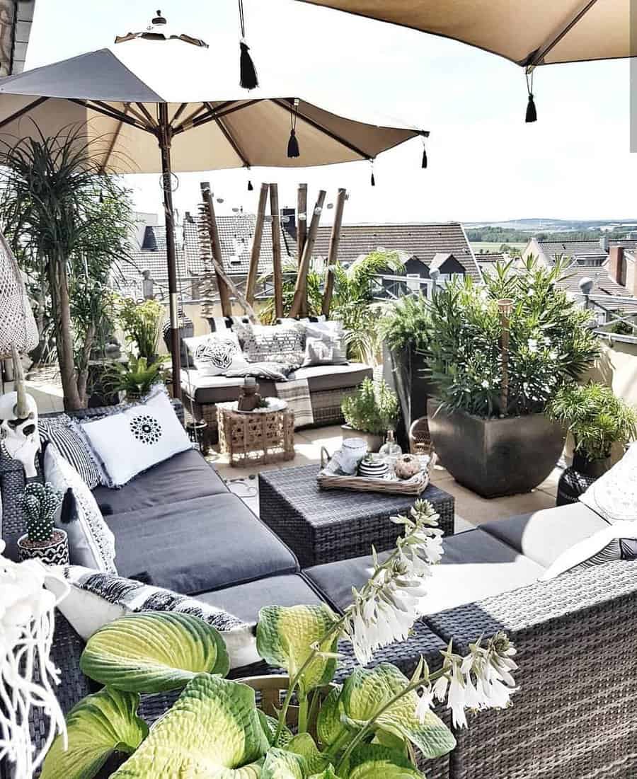 A cozy rooftop terrace with wicker furniture, cushions, large potted plants, and umbrellas, overlooking rooftops and the countryside