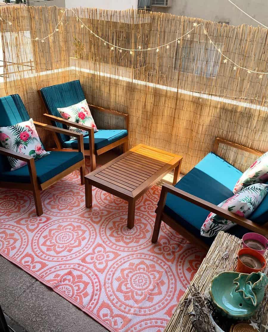 Cozy patio with two wooden chairs, blue cushions, floral pillows, a rug with orange patterns, a wooden table, and string lights above