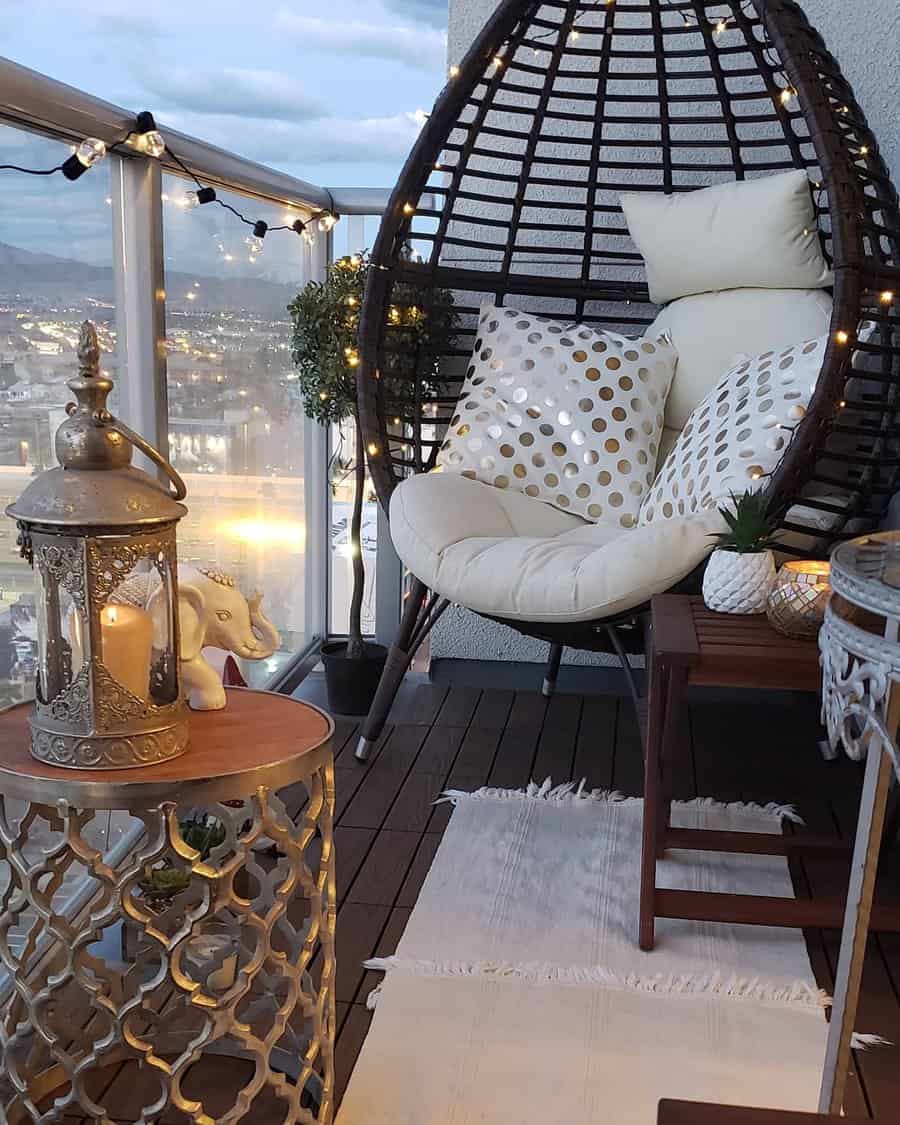 Cozy balcony with a hanging chair, pillows, a small table with a lantern, decor, and string lights, overlooking a cityscape at dusk