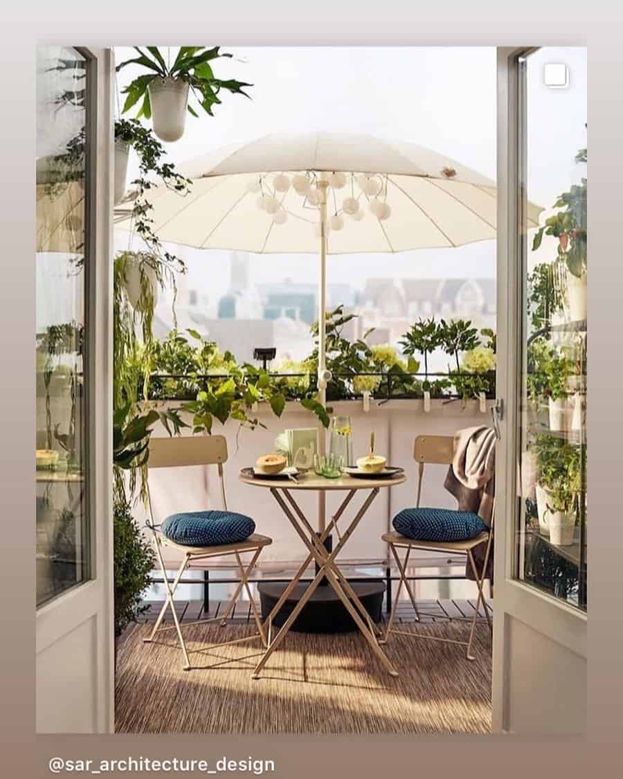 Cozy balcony with a small table set, two chairs, umbrella, and abundant greenery; plates, drinks, and cushions enhance the inviting scene