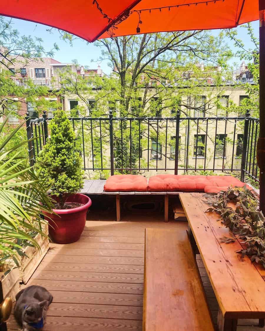 Sunny balcony with orange umbrella, tree view, bench with red cushions, and potted plants on wooden deck