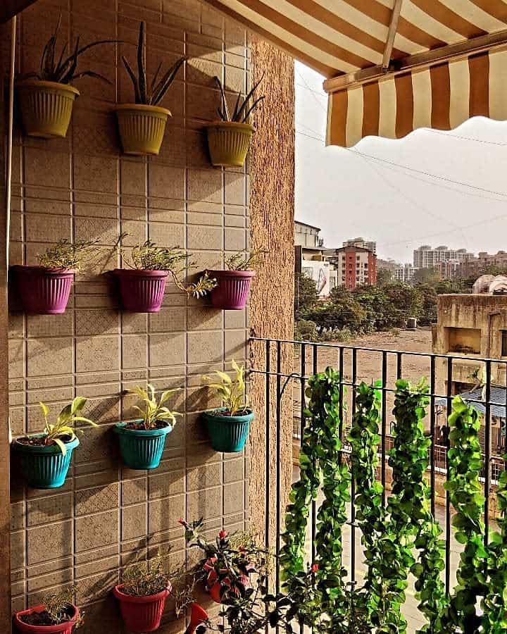 Wall-mounted plants in colorful pots on a balcony with a striped awning, cityscape and greenery visible in the background