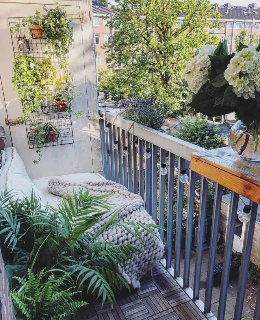 Cozy balcony with plants, a knit blanket on a bench, wall-mounted greenery, string lights, and a view of trees and buildings