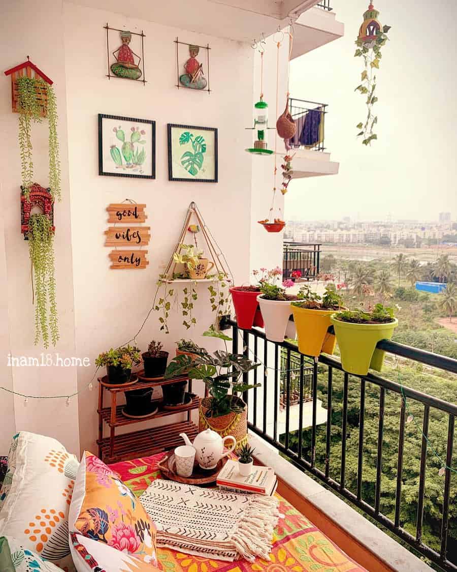 Vibrant balcony with wall art, hanging planters, colorful cushions, potted plants, and a cozy tea setup with scenic city views