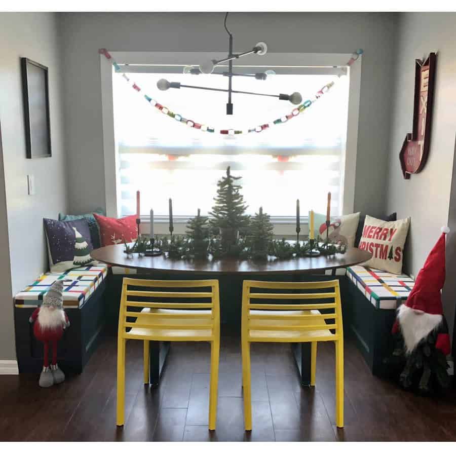 Small dining nook with holiday decor, featuring a round table, yellow chairs, miniature Christmas trees, and festive pillows on benches