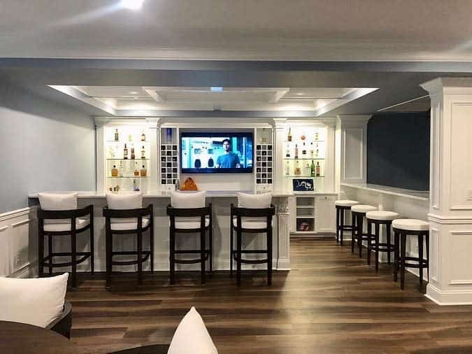 Basement home bar with a sleek, modern design featuring six bar stools, a wall-mounted TV, and shelves stocked with bottles against a wooden floor