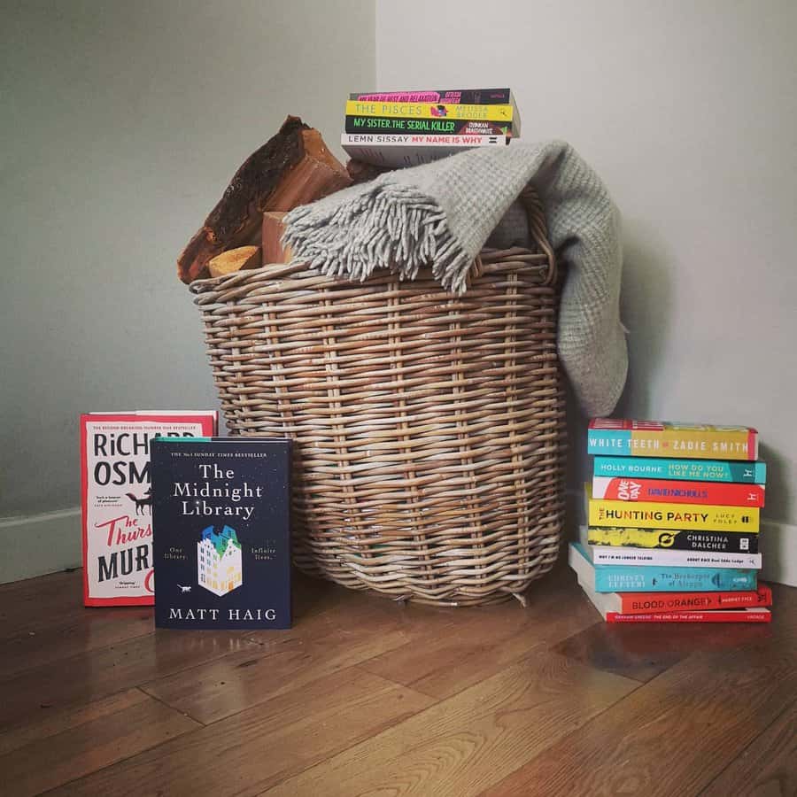  Wicker basket filled with firewood and a cozy blanket, surrounded by stacked books on a wooden floor against a light-colored wall.