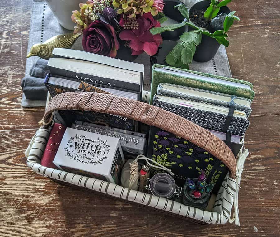 A fabric-lined basket filled with books, journals, tarot cards, and small decorative items, placed on a wooden table with plants and flowers.