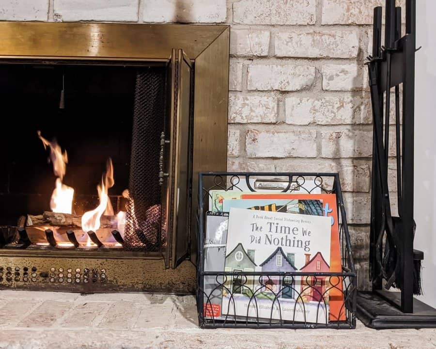 A black wire basket filled with children's books placed beside a cozy fireplace with a brick surround and firewood tools nearby.