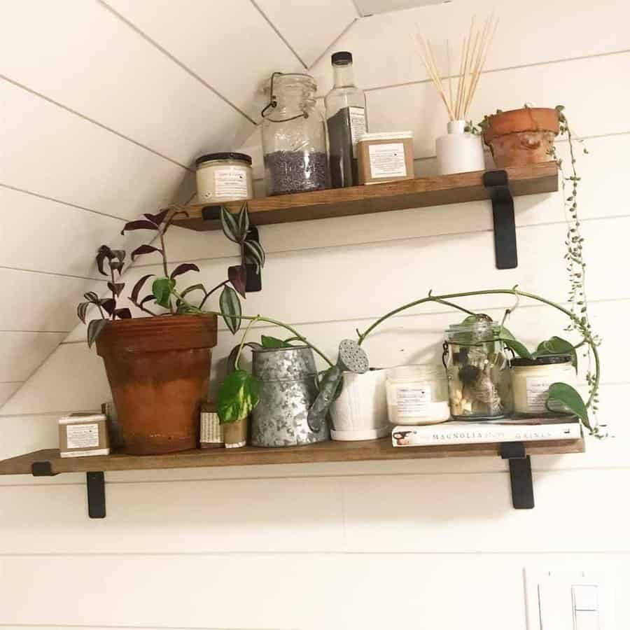 Two wooden shelves with potted plants, candles, a bottle, and small jars against a white shiplap wall