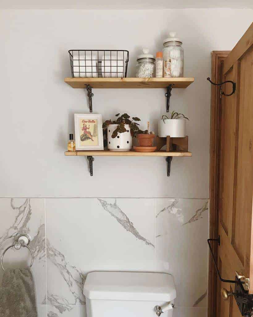 Wooden floating shelves above a toilet with decorative plants, jars, and a wire basket for storage in a modern farmhouse bathroom