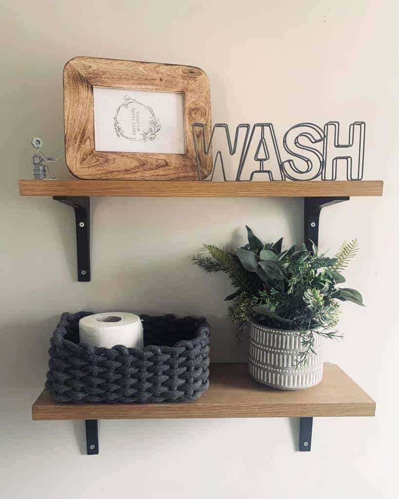 Two wooden shelves with decor: "WASH" sign, framed art, basket with toilet paper, and potted greenery against a beige wall