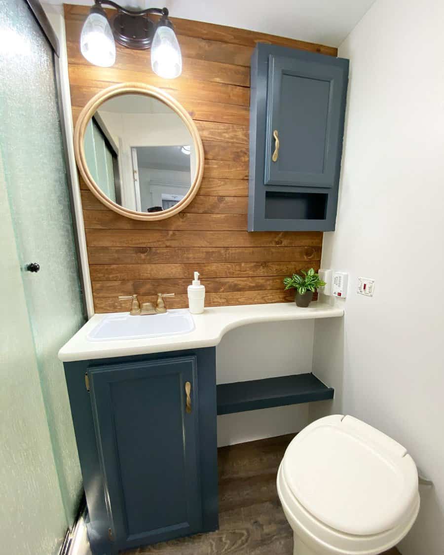 A cozy RV bathroom with a wood accent wall, navy blue cabinetry, a round mirror, and modern fixtures for a stylish, functional look.