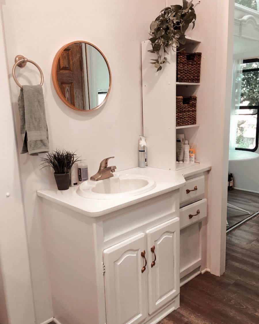 RV bathroom with white cabinetry, a round mirror, woven basket storage, and decorative greenery, creating a cozy and organized space.