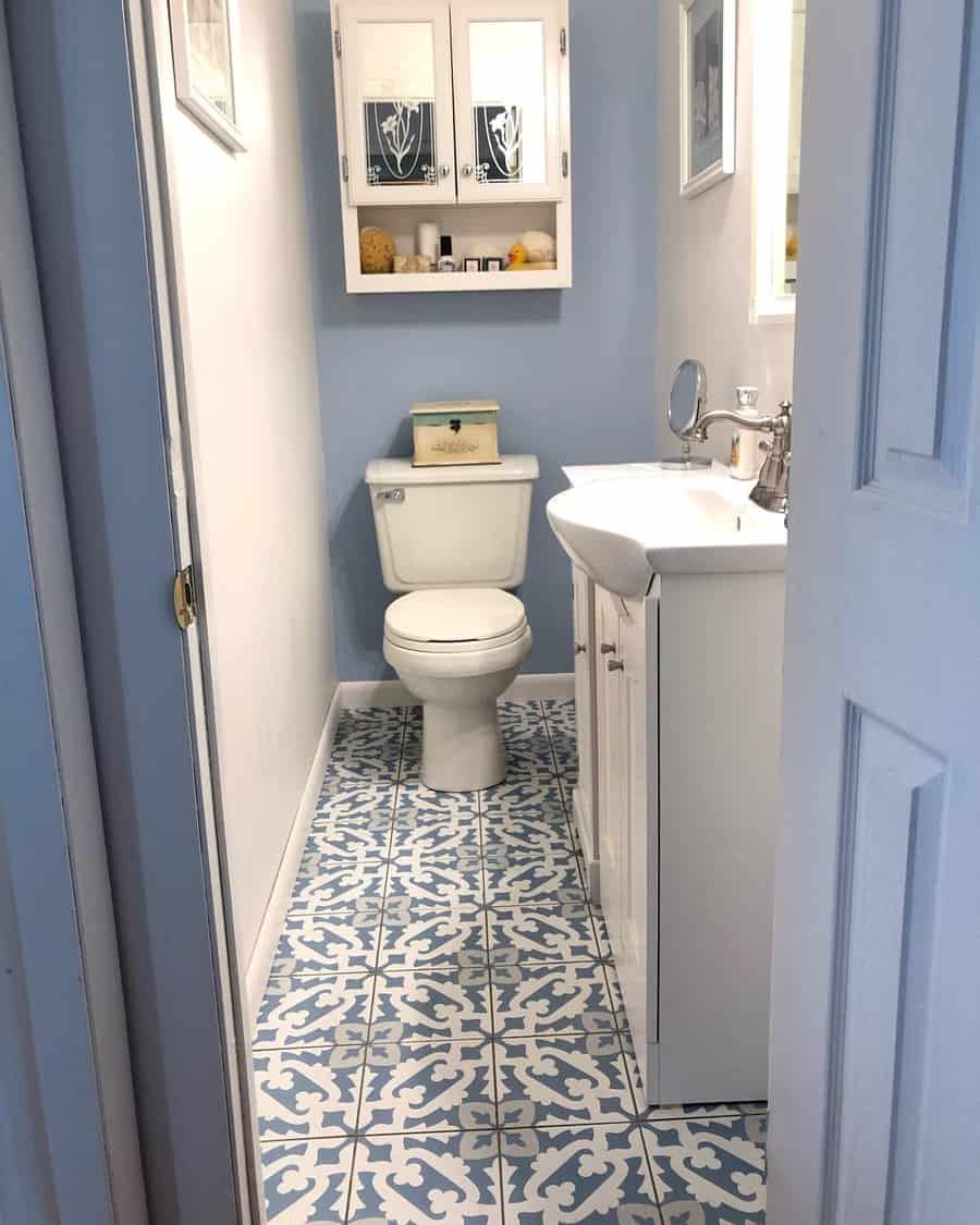 A narrow bathroom with blue and white patterned tile flooring, featuring a white vanity, wall-mounted cabinet, and over-toilet storage