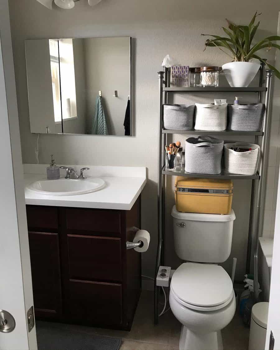 A small bathroom with a sink, mirror, and a storage rack above the toilet holding baskets and a plant, next to a shower area